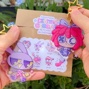 Pode incluir: Dois porta-chaves de acr&iacute;lico transparente com personagens de desenhos animados do The Digital Circus, um coelho roxo e uma boneca de cabelo rosa. Est&atilde;o presos a fechos dourados em forma de estrela sobre um envelope de papel castanho com um autocolante que mostra os nomes Jax, Kinger, Ragatha, Zooble, Caine e Bubble.