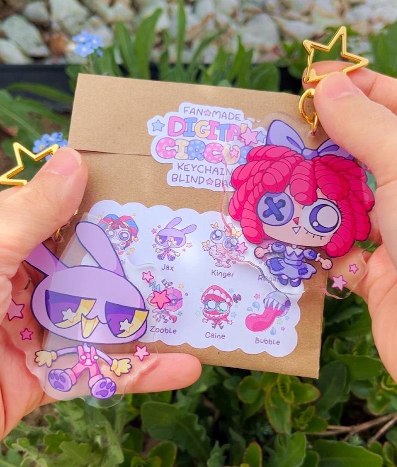 Pode incluir: Dois porta-chaves de acr&iacute;lico transparente com personagens de desenhos animados do The Digital Circus, um coelho roxo e uma boneca de cabelo rosa. Est&atilde;o presos a fechos dourados em forma de estrela sobre um envelope de papel castanho com um autocolante que mostra os nomes Jax, Kinger, Ragatha, Zooble, Caine e Bubble.