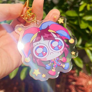 Pode incluir: Um porta-chaves de acr&iacute;lico transparente com uma personagem de desenho animado de olhos grandes, cabelo castanho e um chap&eacute;u de bobo da corte colorido em tons de rosa, azul e amarelo. O design inclui pequenas estrelas amarelas e um fecho met&aacute;lico dourado em forma de estrela.