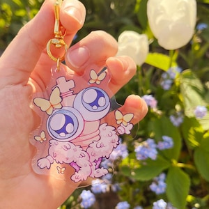 Pode incluir: Um porta-chaves de acr&iacute;lico transparente com uma personagem de desenho animado com dois grandes olhos roxos, um corpo rosa felpudo e pequenas borboletas amarelas. O pendente est&aacute; preso a um fecho lagosta e anel de metal dourado.