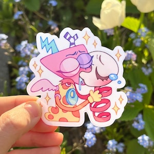 Op de afbeelding: Een kleurrijke sticker met twee stripfiguren die elkaar omarmen. De ene figuur is roze met een blauwe kroon, de andere wit met een blauwe traan. De sticker heeft een witte rand en gouden sterren.