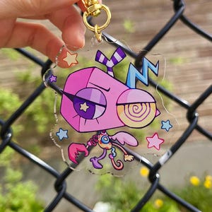 Pode incluir: Um porta-chaves de acr&iacute;lico transparente com uma personagem de desenho animado com uma cabe&ccedil;a geom&eacute;trica cor-de-rosa, um olho em forma de estrela e um olho em espiral. A personagem tem um corpo pequeno com m&atilde;os em garra e asas, rodeada de estrelas.