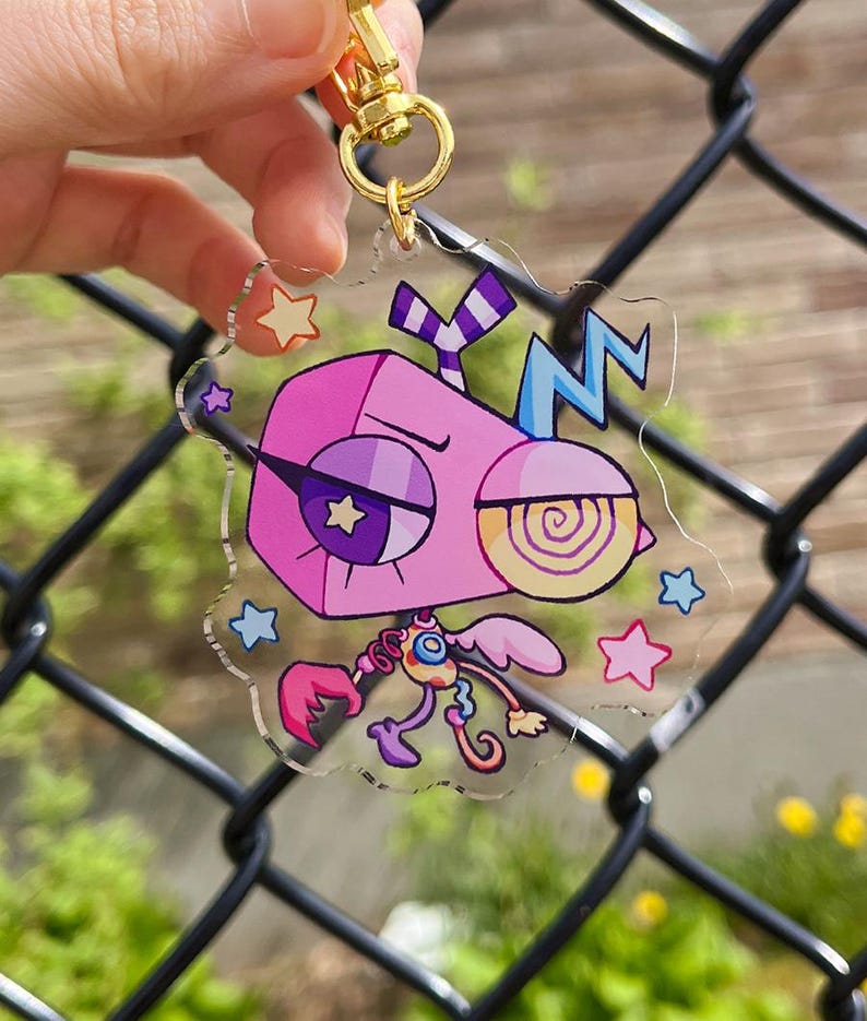 Pode incluir: Um porta-chaves de acr&iacute;lico transparente com uma personagem de desenho animado com uma cabe&ccedil;a geom&eacute;trica cor-de-rosa, um olho em forma de estrela e um olho em espiral. A personagem tem um corpo pequeno com m&atilde;os em garra e asas, rodeada de estrelas.