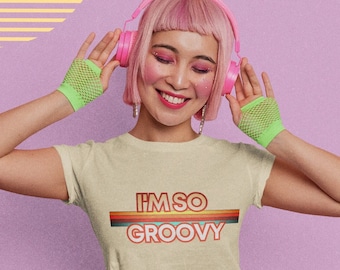 May include: A woman with pink hair wearing a light beige t-shirt with the text "I'M SO GROOVY" in rainbow colors. She is wearing headphones and green mesh fingerless gloves.