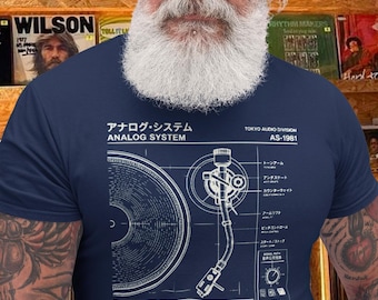 Puede incluir: Camiseta azul marino con un plano detallado de una unidad de reproducción de vinilo, que incluye un brazo fonocaptor y un contrapeso. La camiseta tiene texto japonés y las palabras "ANALOG SYSTEM" y "VINYL PLAYBACK UNIT". El fondo muestra una colección de discos de vinilo.