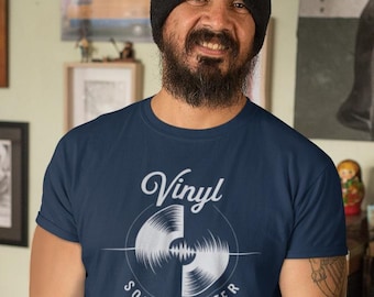 Puede incluir: Una persona con una camiseta azul marino con el texto "Vinyl Sounds Better" y un gráfico de un disco de vinilo. La persona sostiene una guitarra eléctrica con un acabado marrón y naranja. El fondo incluye obras de arte enmarcadas y un letrero de "Fans Parking".