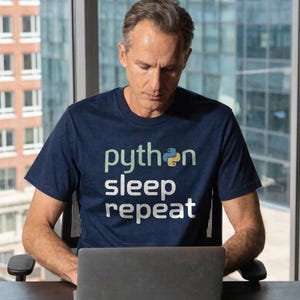 May include: A navy blue t-shirt with the words "python sleep repeat" in white and light green text. The shirt features a Python logo. The person is sitting at a desk with a laptop.
