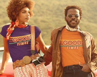 May include: Two people wearing vintage-style t-shirts with the text "Good Times" in rainbow lettering. The woman is wearing bell-bottom pants with a floral pattern and a purple shirt. The man is wearing a brown leather jacket and blue jeans. They are standing in front of a red car.