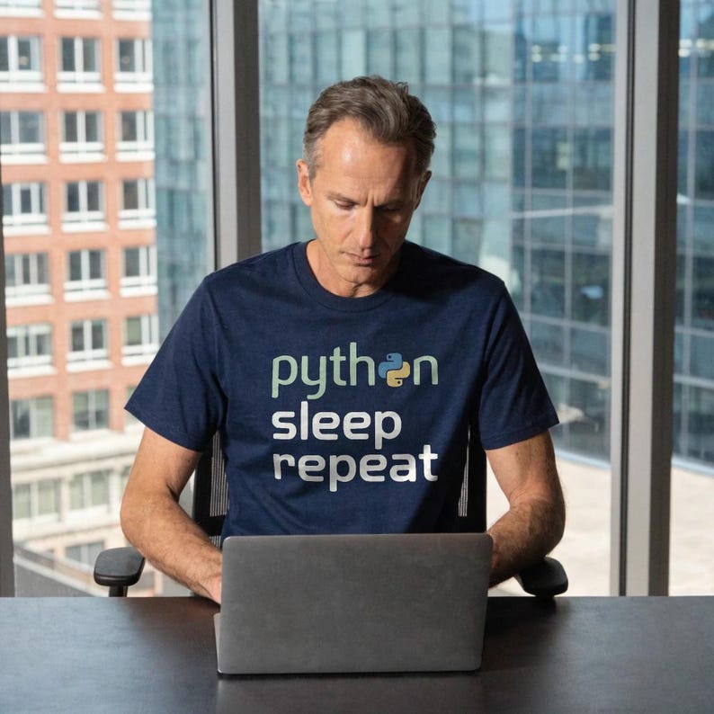May include: A navy blue t-shirt with the words "python sleep repeat" in white and light green text. The shirt features a Python logo. The person is sitting at a desk with a laptop.