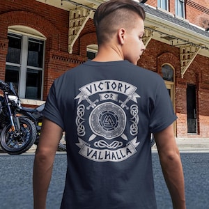 Könnte beinhalten: Dunkelblaues T-Shirt mit weißem Aufdruck auf dem Rücken. Der Aufdruck zeigt "VICTORY OR VALHALLA" mit Schild, Schwertern und Wikingerknoten. Im Hintergrund ist ein Motorrad zu sehen.
