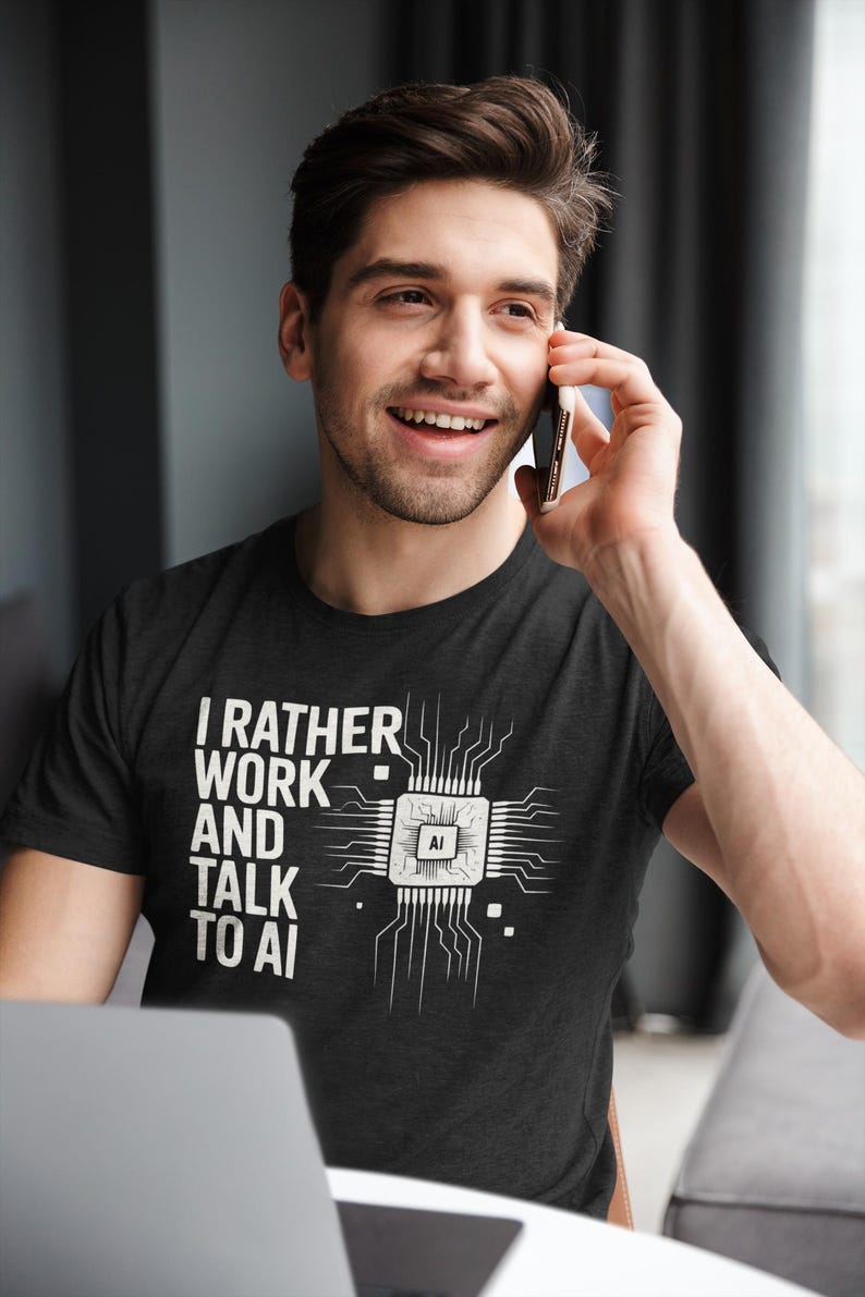 May include: A dark gray t-shirt with the text "I RATHER WORK AND TALK TO AI" and a white graphic of a computer chip. A person is wearing the shirt and talking on a smartphone.
