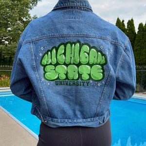 Puede incluir: Una chaqueta vaquera azul con un gráfico verde y blanco de "Michigan State University" pintado en la espalda.