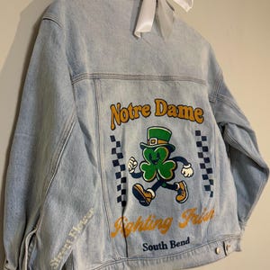 Puede incluir: Chaqueta vaquera azul claro con un diseño de duende y trébol, con las palabras "Notre Dame Fighting Irish South Bend". La manga tiene las palabras "Street Flower".