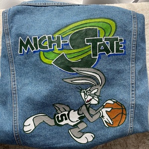Puede incluir: Una chaqueta vaquera azul con un diseño de dibujos animados verde y amarillo de Bugs Bunny jugando al baloncesto. El texto "MICH-TATE" está escrito en la chaqueta.