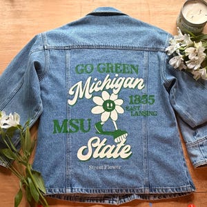 Puede incluir: Chaqueta vaquera azul claro con texto gráfico verde y blanco y un diseño de flor en la espalda. El texto dice "GO GREEN Michigan 1855 East Lansing MSU State Street Flower". La chaqueta tiene mangas largas y cierre de botones.