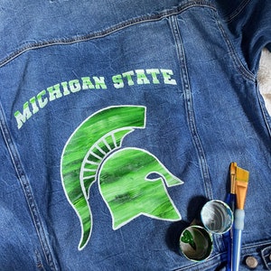 Puede incluir: Una chaqueta vaquera azul con un casco espartano pintado en verde y blanco y las palabras "Michigan State" en la espalda. La chaqueta está sobre una superficie blanca con pinceles y botes de pintura.