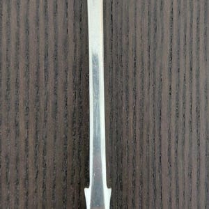 May include: A silver-toned metal spoon featuring a long, slender handle with a flared, rounded end. The handle is engraved with the cursive letters 'W' and 'M'. The bowl of the spoon is oval-shaped and polished to a reflective, mirror-like finish.