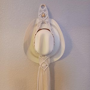 May include: A white cowboy hat hangs from a macrame hat hanger. The hat has a wide brim with a perforated design. The macrame is cream-colored and features a wooden ring at the top and a tassel at the bottom. The hat and hanger are mounted on a textured wall.