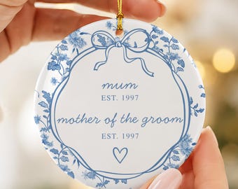 May include: A round white ceramic ornament featuring a blue floral border and a central blue ribbon graphic. The text reads "mum EST. 1997" and "mother of the groom EST. 1997" with a small heart icon below. It hangs from a thin gold string.