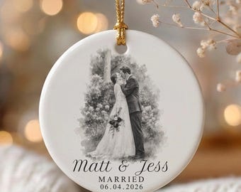 May include: A round white ceramic ornament hanging from a gold cord. The center features a black and white sketch of a couple embracing in wedding attire. Below the image, the text reads Matt & Jess, MARRIED, and the date 06.04.2026.