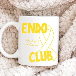 May include: White ceramic mug featuring the words "ENDO CLUB" in yellow, with a yellow ribbon graphic and the text "it's not just a bad period". The mug is displayed on a soft, textured surface.