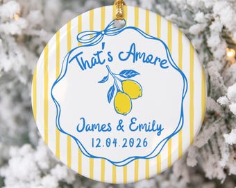 May include: A round ceramic ornament featuring a yellow and white vertical striped pattern. The center displays a blue scalloped frame with a lemon illustration, the text That's Amore, the names James & Emily, and the date 12.04.2026 in blue.
