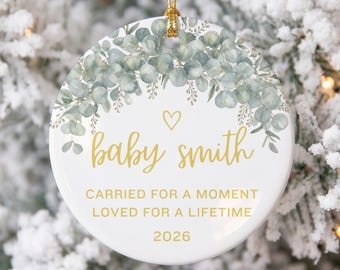 May include: A round white ceramic ornament featuring a watercolor eucalyptus wreath at the top, a gold heart, and gold text that reads baby smith, CARRIED FOR A MOMENT LOVED FOR A LIFETIME, and 2026. It hangs from a gold string.