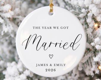 May include: A round white ceramic ornament featuring the black printed text THE YEAR WE GOT Married, a small heart icon, the names JAMES & EMILY, and the year 2026. The ornament is suspended by a thin gold cord through a hole at the top center.