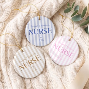 May include: Three round ceramic ornaments with gold hanging strings. Each features vertical stripes and the text "The year I became a NURSE" with a heart and the year "2026". One is light blue, one is light pink, and one is light tan.