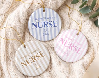 May include: Three round ceramic ornaments with gold hanging strings. Each features vertical stripes and the text "The year I became a NURSE" with a heart and the year "2026". One is light blue, one is light pink, and one is light tan.