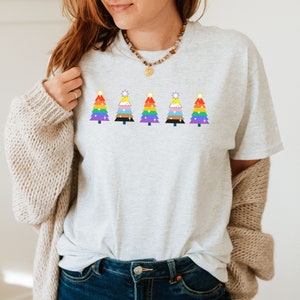 Może przedstawiać: Szary T-shirt z rzędem pięciu tęczowych choinek. Każda choinka ma gwiazdę na szczycie i jest ozdobiona białym brokatem.