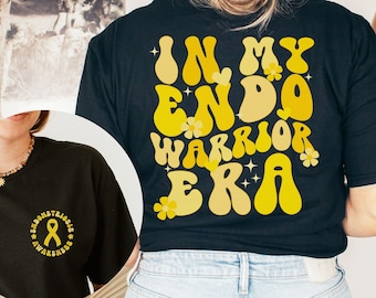 May include: Black t-shirt with yellow text that reads "In My Endo Warrior Era". The text is in a retro font style with flowers and stars.