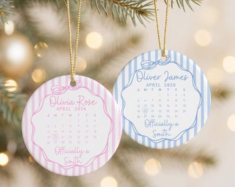 May include: Two round ceramic ornaments with gold hanging strings. One features pink vertical stripes, the name Olivia Rose, April 2026 calendar, and the text Officially a Smith. The other has blue stripes, Oliver James, and the same text.