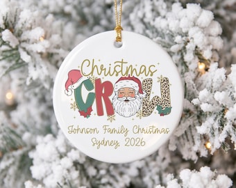 May include: A round white ceramic Christmas ornament with a gold string. The text reads Christmas Crew, Johnson Family Christmas, Sydney, 2026. The design features a Santa Claus illustration, red and green letters, and leopard print accents.