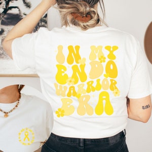 May include: White t-shirt with a yellow graphic that says "In My Endo Warrior Era" with yellow flowers around the text.