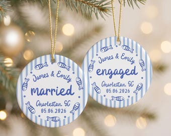 May include: Two round ceramic ornaments with a blue and white vertical striped pattern. Each features blue text reading James & Emily, either married or engaged, Charleston, SC, and 05.06.2026, surrounded by small blue fish illustrations and gold hanging cords.