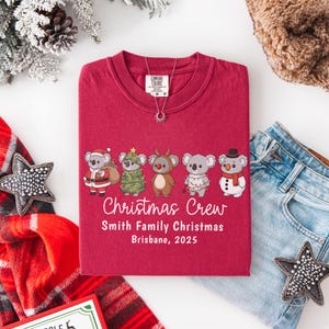 May include: A maroon t-shirt featuring a graphic of six koalas dressed for Christmas. The text "Christmas Crew" is below the koalas, with "Smith Family Christmas Brisbane, 2025" underneath. A silver necklace is on the shirt.