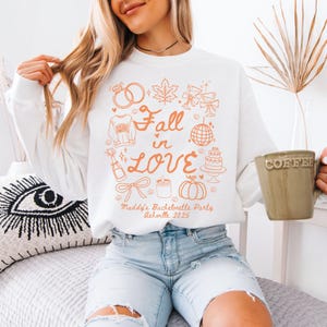 May include: White sweatshirt featuring an orange graphic with the words "Fall in Love" and illustrations of rings, leaves, champagne, a disco ball, cake, and a pumpkin. The text "Maddy's Bachelorette Party Asheville 2025" is also printed.
