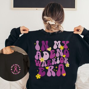 May include: Black sweatshirt with a purple and pink graphic that says "In My Adeno Warrior Era" with yellow flowers and hearts. The back of the sweatshirt has a purple ribbon with the text "Adeno Awareness" in a circle.