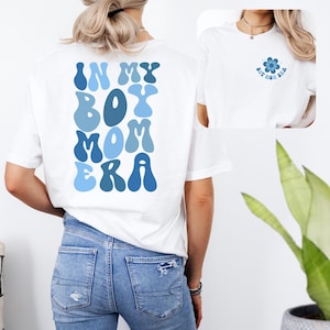 Könnte beinhalten: Weißes T-Shirt mit blauem Text, der auf der Rückseite "In My Boy Mom Era" lautet. Die Vorderseite des Shirts hat eine kleine blaue Blume mit dem gleichen Text darin.