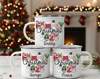 Könnte beinhalten: Drei weiße Emaille-Tassen mit schwarzen Rändern sind gestapelt. Jede Tasse hat einen anderen Namen: Daddy, George und Mama. Die Tassen zeigen den Text "Family Christmas 2025" mit festlichen Illustrationen von Rentieren, Schneemännern und Fäustlingen.