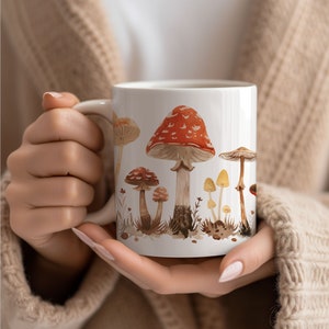 Puede incluir: Taza de cerámica blanca con una ilustración de acuarela de varios hongos en tonos de rojo, marrón y amarillo. Los hongos se representan en un entorno de bosque con follaje verde.