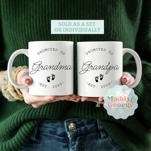 May include: Two white ceramic mugs with black text and a footprint design. The mug on the left says "Promoted to Grandma - Est. 2025" and the mug on the right says "Promoted to Grandpa - Est. 2025".