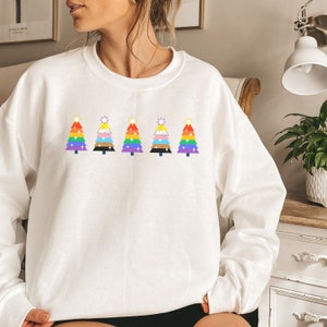 May include: White sweatshirt with five rainbow-coloured Christmas tree designs. Each tree features a different pride flag design, including the LGBTQ+ rainbow flag, transgender flag, and other pride flags, topped with a star.