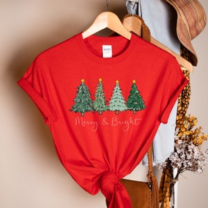 以下が含まれることがあります： 緑のクリスマスツリー4本が金色に輝く星と光で飾られた赤いTシャツ。ツリーの下には「Merry & Bright」の文字がプリントされています。