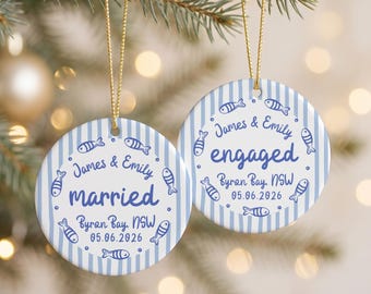 May include: Two round ceramic ornaments with a light blue and white vertical striped pattern. Each features blue text reading James & Emily, Byron Bay, NSW, 05.06.2026, with one saying married and the other engaged, surrounded by small blue fish icons.