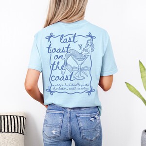 Peut inclure: T-shirt bleu clair avec un motif de sirène dans un verre à cocktail et le texte "last toast on the coast". Le texte "maddy's bachelorette party, Charleston, South Carolina" est également présent.
