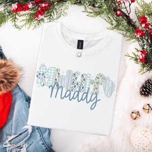 以下が含まれることがあります： 青い筆記体で「Maddye」という名前が書かれた白いTシャツ。名前はギンガムチェックや花柄など、青と白の模様で装飾されています。Tシャツは折りたたまれ、お祝いの装飾に囲まれています。