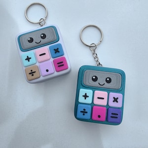 May include: Two calculator keychains with smiling faces on the screens. The left keychain is white with pastel buttons in blue, pink, and tan. The right keychain is teal with buttons in light blue, pink, purple, and dark pink. Both have metal rings.