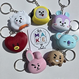 May include: A collection of seven character-themed keychains featuring various shapes and colors, including a white cloud, yellow dog, purple mouse, red heart, light blue koala, pink rabbit, and tan cookie, surrounding a square card with the text We MAKE DESIGN LAB.